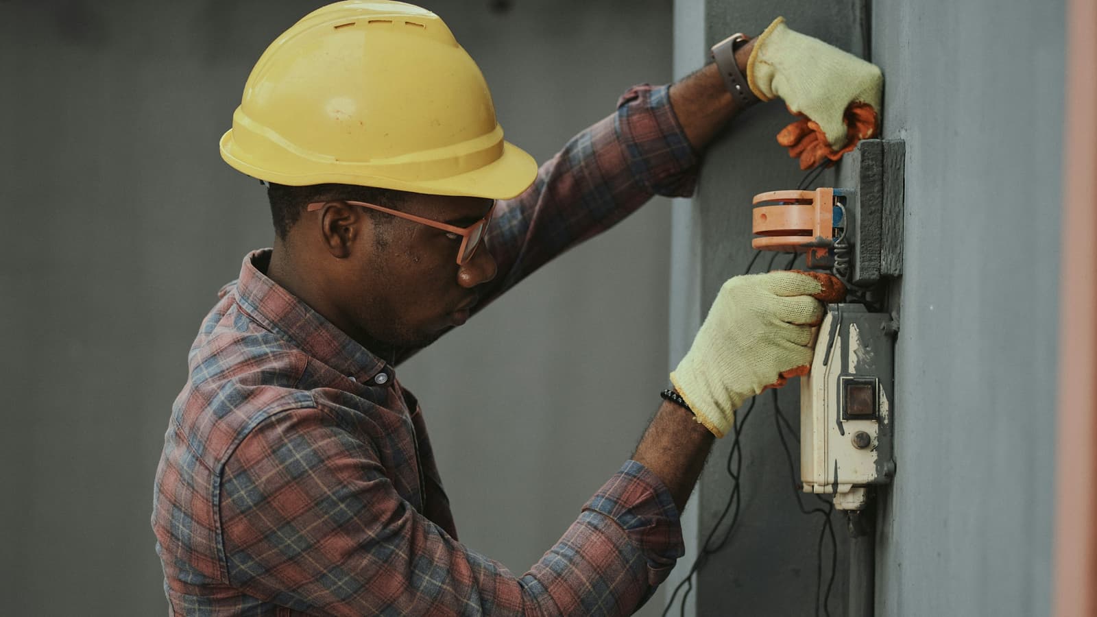 Electrician in a yellow hard hat and gloves working on a wall-mounted electrical box